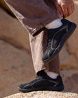Person adjusting DANNTO Aero N1 black barefoot shoes while standing on rocks outdoors