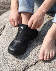 Dannto Frond - Non-Slip Zero Drop Barefoot Sneakers black shown being put on while sitting on a rock outdoors