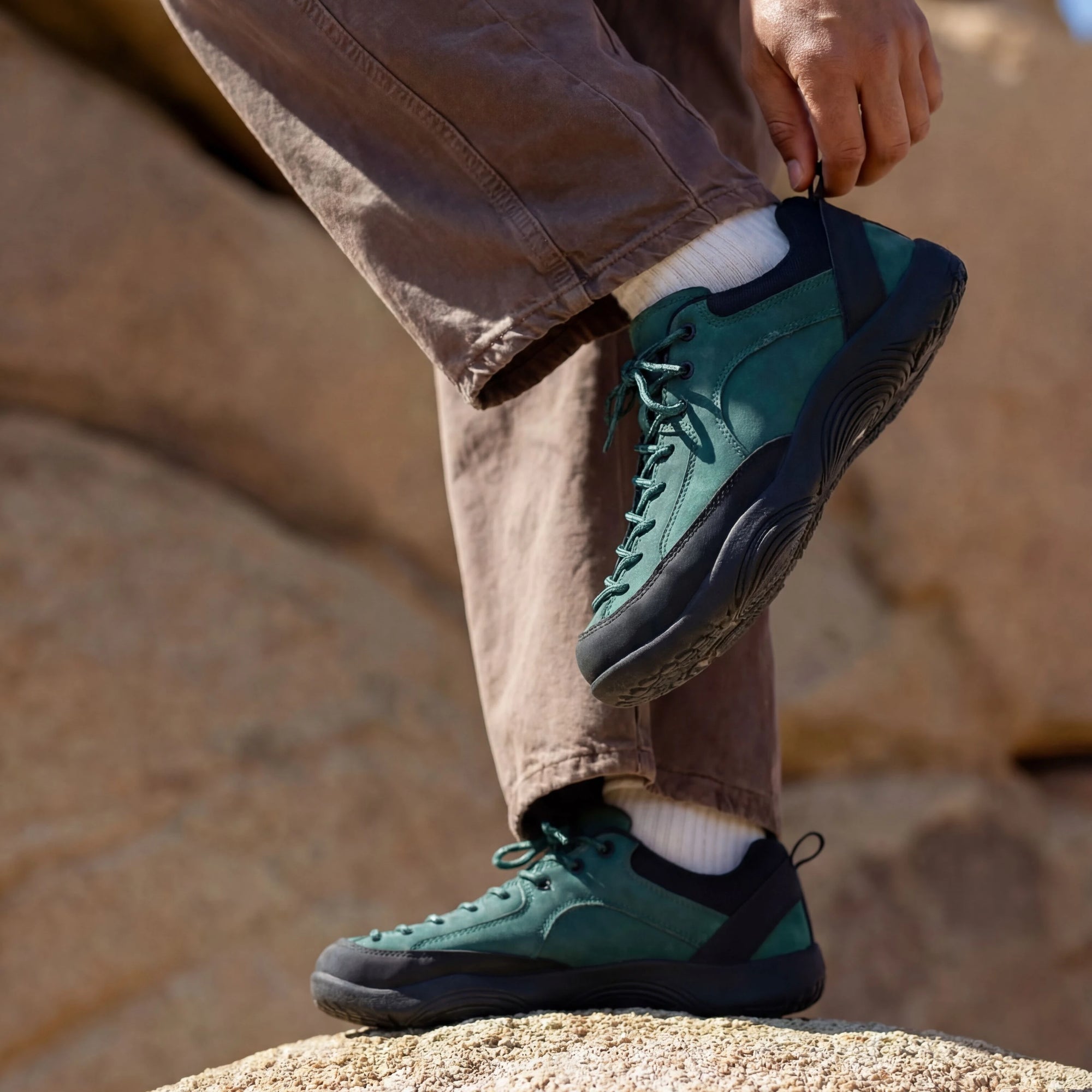 Person adjusting DANNTO Veld I green   barefoot shoes while standing on rocks outdoors