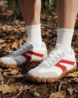 DANNTO Purus Men's and Women's Wide Toe Box Barefoot Shoes in White Worn While Standing on a Leaf-Covered Forest Floor.