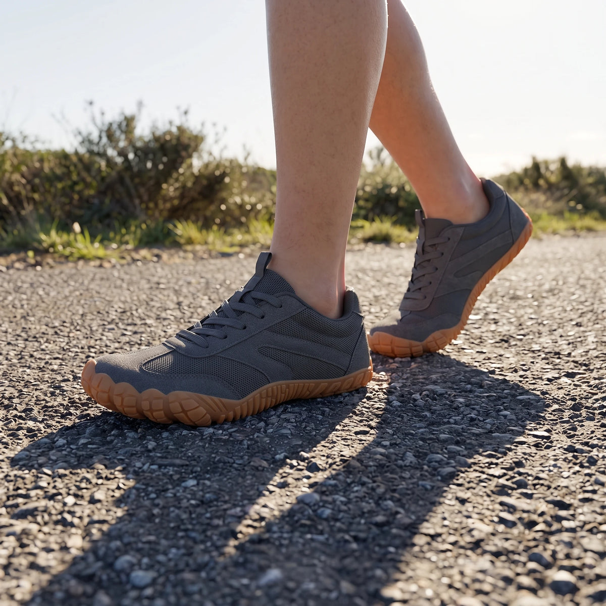 DANNTO Men's and Women's Wide Toe Box Barefoot Shoes in Gray Worn While Walking on a Sunlit Gravel Path.