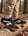DANNTO Purus Men's and Women's Wide Toe Box Barefoot Shoes in Black Worn While Walking Across a Fallen Log in a Forest.