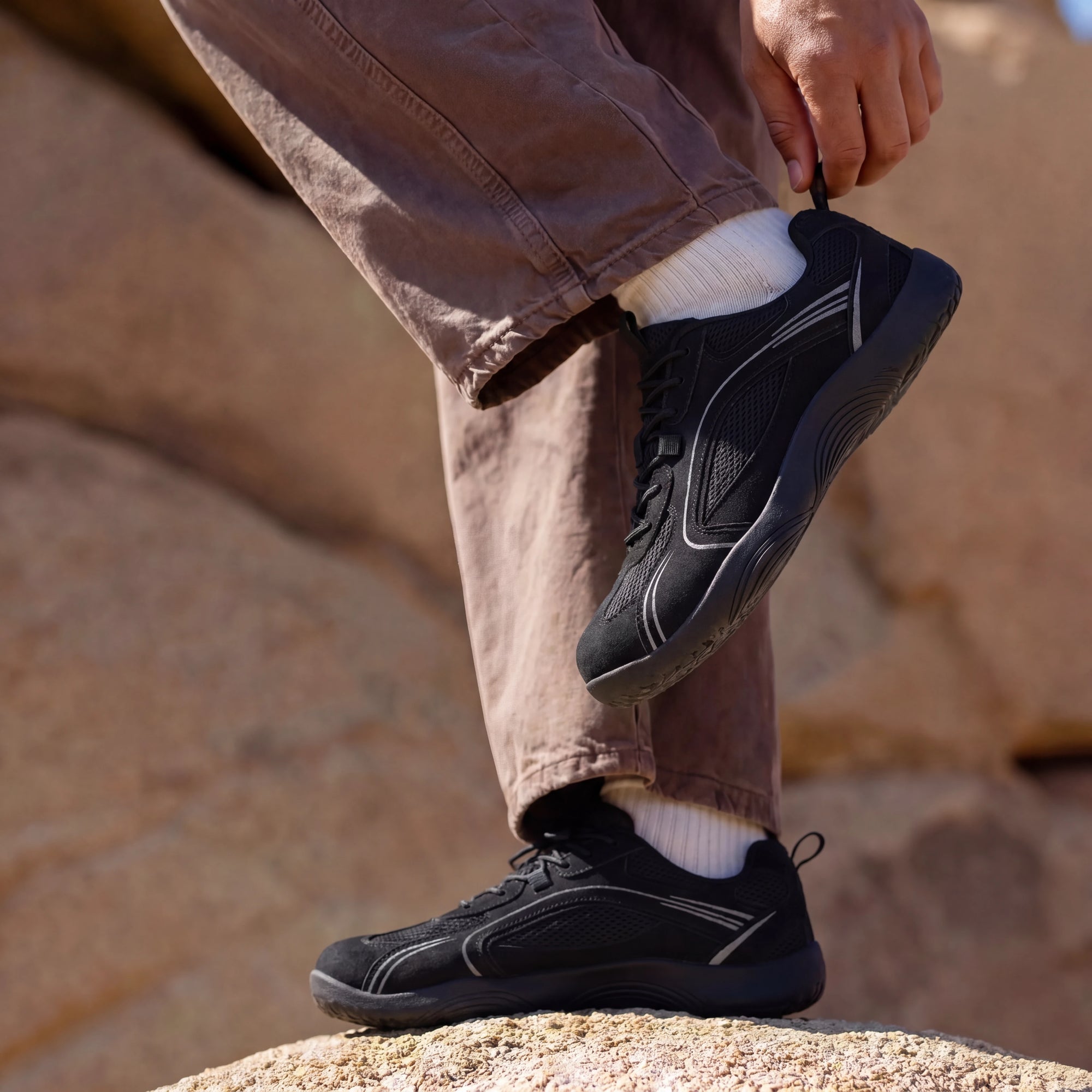 Person adjusting DANNTO Aero N1 black barefoot shoes while standing on rocks outdoors