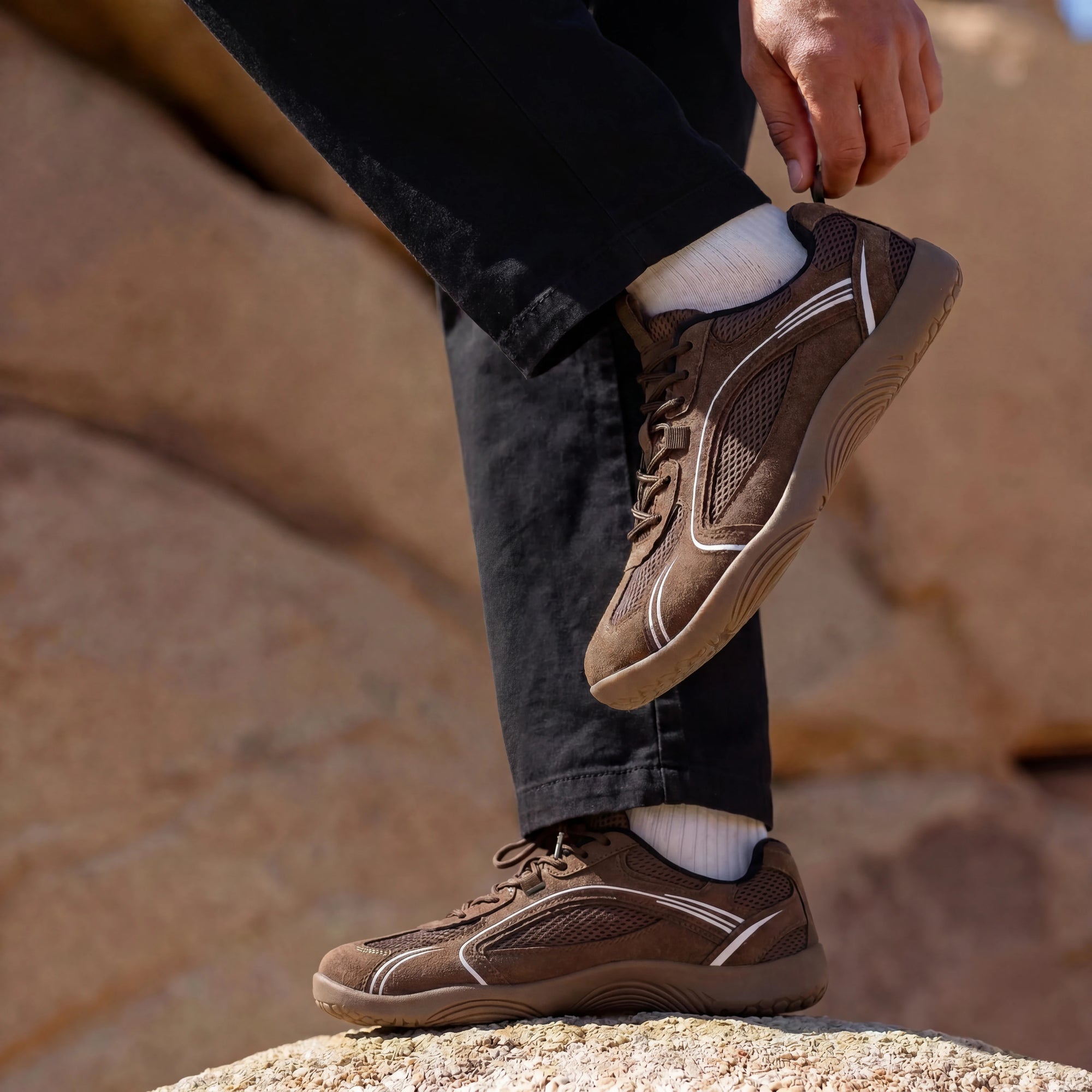 Person adjusting DANNTO Aero N1 brown barefoot shoes while standing on rocks outdoors